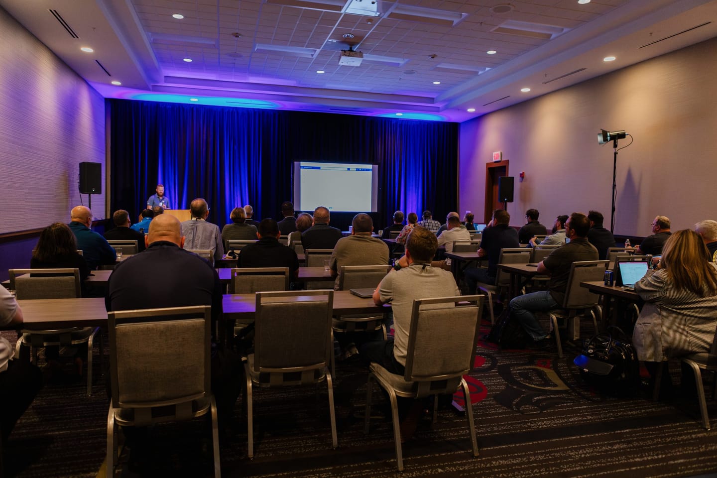 A full session room at the ACCIS Fall Conference 2025, with members at tables facing a speaker at the podium under blue stage lighting.